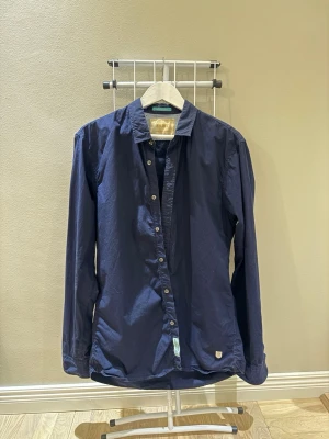 Mörkblå skjorta från Scotch & Soda - Snygg mörkblå skjorta från Scotch & Soda med klassisk krage och knäppning framtill. Skjortan har långa ärmar och en liten läderbricka vid nederkanten. Perfekt för en stilren look.