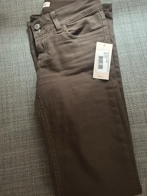 Bruna jeans från Nelly - Snygga bruna jeans från Nelly med utsvängda ben och klassisk femficksmodell. Jeansen har dekorativ söm på bakfickan och silverfärgade knappar. Materialet känns mjukt och stretchigt, perfekt för en avslappnad look.