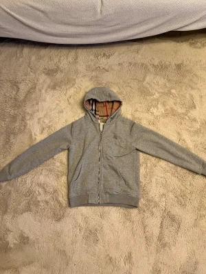 Grå zip hoodie från Burberry - Snygg grå hoodie med dragkedja från Burberry. Klassisk huva med det ikoniska rutmönstret på insidan och broderad logga på bröstet. Tillverkad i mjukt bomullsmaterial och har två fickor framtill. Perfekt för en avslappnad och stilren look.