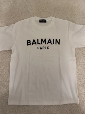 Vit Balmain Paris t-shirt - Vit t-shirt från Balmain Paris med svart tryck framtill. Klassisk rund halsringning och korta ärmar. Tillverkad i mjuk bomull för en skön och stilren look.