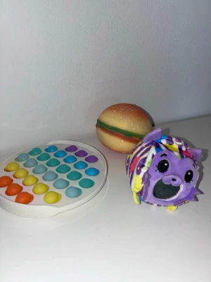 Fidgetleksaker och squishy-bollar - Säljer ett kit med fidgetleksaker: en rund pop it i regnbågsfärger, en squishy-boll som ser ut som en hamburgare och en lila mjukisdjur squishy boll med glitter. Perfekt för dig som gillar att pilla och stressa ner.