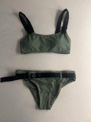 Olivgrön bikini med svarta detaljer - Snygg olivgrön bikini med svarta axelband och ett coolt svart bälte med metallspänne på underdelen. Sportig topp med breda band och trendig look. Perfekt för stranden eller poolen. OANVÄND!😅