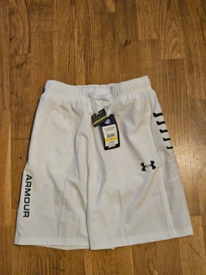 Vita sportiga shorts från Under Armour - Snygga vita shorts från Under Armour med svart logga och text på ena benet. De har elastisk midja med snörning och är tillverkade i ett lätt och funktionellt material som passar perfekt för träning. Svarta detaljer på sidorna ger en sportig look.