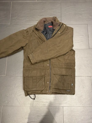 Brun vintage parkajacka från Fjällräven g-1000 - Klassisk brun vintage parkajacka från fjällräven med flera stora fickor framtill, hög krage och dragkedja. Jackan har justerbar nederkant med snörning och är tillverkad i slitstarkt material, perfekt för utomhusaktiviteter. Fodrad insida för extra komfort. Knappt använd, väldigt fint skick. Nypris 6500kr, Storlek M. Säljer den för 2000kr, pris kan diskuteras. 