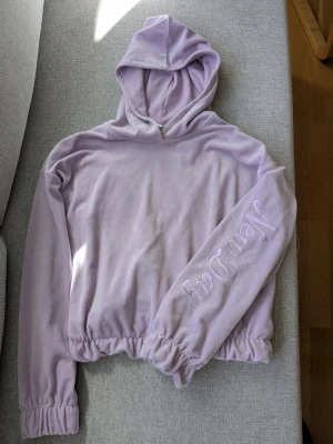 Ljuslila hoodie med text New Day - Mysig ljuslila hoodie i mjukt velourmaterial med huva och resår i midja och ärmslut. På ena ärmen står det 'New Day' i stora bokstäver med små glittriga detaljer runt texten. Perfekt för en chill och trendig look.