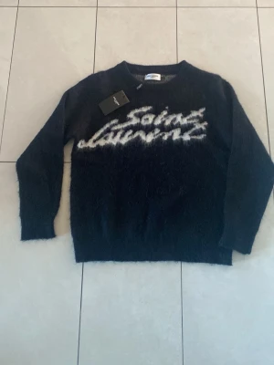 Svart Saint Laurent stickad tröja - Snygg svart stickad tröja från Saint Laurent med vit logotyp i graffiti-stil över bröstet. Tröjan har rund halsringning, långa ärmar och är gjord i ett fluffigt, mjukt material som ger en cool och trendig look.