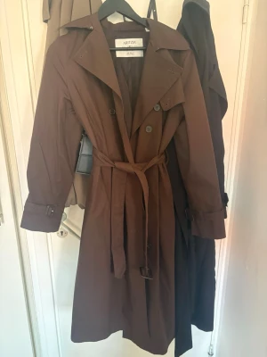 Brun trenchcoat från Aritzia - Snygg brun trenchcoat från Aritzia med klassisk dubbelknäppning, midjebälte och justerbara spännen vid ärmsluten. Jackan har bred krage och raka ärmar, perfekt för en stilren look. Materialet är slätt och ger en modern känsla.