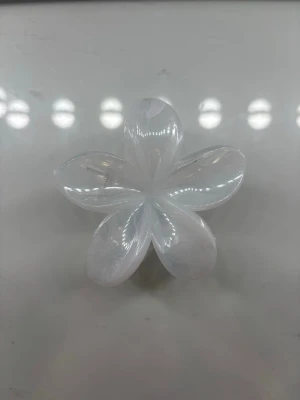 Vit blomformad hårklämma - Säljer en söt hårklämma i form av en blomma, tillverkad i genomskinlig vit plast med pärlemorskimrande yta. Klämman har fem kronblad och en guldfärgad fjäder. Perfekt för att ge frisyren en extra touch.