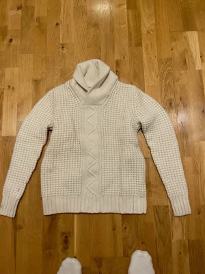Valient Vit stickad polotröja med mönster stl M - Mysig vit stickad polotröja med grov struktur och kabelstickat mönster framtill. Tröjan har lång ärm, bred ribbad mudd vid ärmslut och nederkant samt en hög, lös polokrage. Perfekt för kalla dagar.