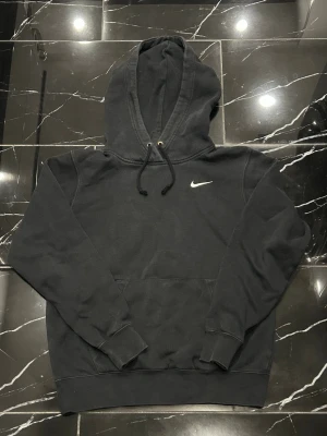 Svart Nike hoodie med logga - Svart hoodie från Nike med klassisk vit swoosh-logga på bröstet. Tröjan har huva med dragsko och en stor magficka. Perfekt för en avslappnad och sportig stil. Storlek xs