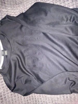 Svart sweatshirt från Hugo Boss - Svart sweatshirt med rund hals från Hugo Boss. Tröjan har ett stort, ton-i-ton broderat BOSS-logo på bröstet och är tillverkad i mjukt material. Perfekt för en stilren och avslappnad look.