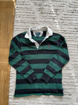 Randig rugbytröja från Polo Ralph Lauren - Grön och svart randig rugbytröja från Polo Ralph Lauren med vit krage och broderad röd logga på bröstet. Långärmad modell i bomull med knappar vid halsen. Perfekt för en sportig och avslappnad stil. storlek M i barn 