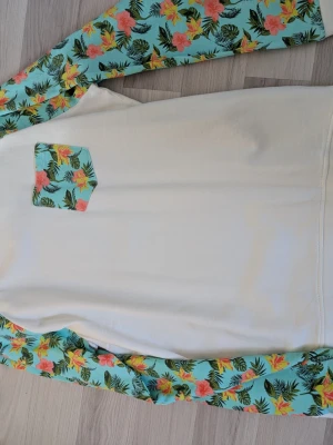 Vit tröja med blommiga ärmar Lager 157 DOG - Snygg vit tröja från Lager 157 DOG med blommiga ärmar och matchande bröstficka. Ärmarna har tropiskt mönster i turkos, rosa, gul och grön. Perfekt för dig som vill sticka ut med en unik och färgglad stil. Mjuk bomullskänsla och avslappnad passform.