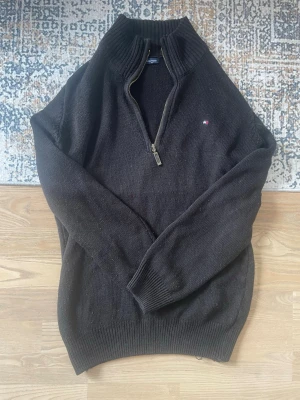 TOMMY HILFIGER Svart stickad tröja  - Svart stickad tröja från Tommy Hilfiger med halv dragkedja och ståkrage. Liten broderad logga på bröstet. Tröjan har långa ärmar och ribbade muddar vid ärmslut och nederkant. Perfekt för lager-på-lager-stil.