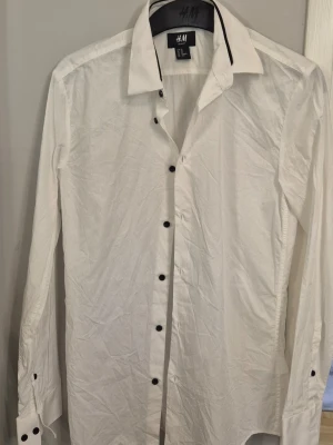 Vit slim fit skjorta från H&M - Klassisk vit skjorta från H&M i slim fit-modell. Skjortan har svarta knappar och en snygg detalj med svart kant på kragen. Tillverkad i bomull och har långa ärmar med knappslå vid manschetterna. Perfekt för en stilren look.