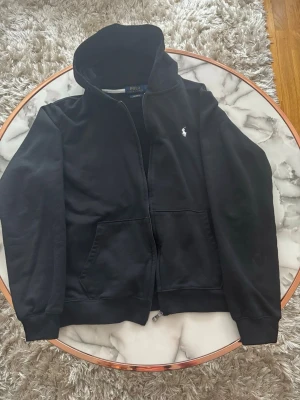Svart hoodie från Polo Ralph Lauren - Svart hoodie med dragkedja från Polo Ralph Lauren. Klassisk design med vit broderad logga på bröstet, kängurufickor och ribbade muddar. Mjuk bomullskvalitet och skön huva. Perfekt för en avslappnad och stilren look.