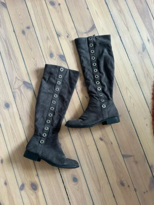 Grå höga stövlar med detaljer  - Säljer ett par gråa höga stövlar i mockaimitation med stora silverfärgade nitar längs utsidan. De har låg klack och rundad tå, vilket ger en cool och edgy look. Perfekta för dig som vill sticka ut lite extra.