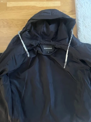 Svart bomberjacka från Emporio Armani - Snygg svart bomberjacka från Emporio Armani med huva och dragsko. Jackan är tillverkad i micro twill av 100% polyester och har en stilren design med innerficka och dragkedja. Perfekt för dig som vill ha en klassisk och trendig look.