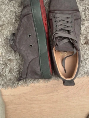 Mörkgråa sneakers i mocka - Snygga mörkgråa sneakers från Christian Louboutin i mjuk mocka. Skorna har svarta sulor med röd detalj på sidan, matchande grå skosnören och diskret branding på insidan. Perfekta för dig som gillar stilrena och exklusiva sneakers.