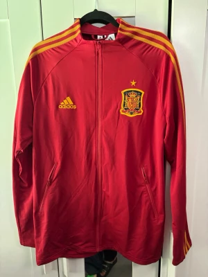 Röd Adidas Spanien träningsjacka - Röd träningsjacka från Adidas med Spaniens landslagsmärke på bröstet och gula detaljer. Jackan har hel dragkedja, två fickor och gula ränder längs ärmarna. Perfekt för fotbollsfans som vill representera Spanien. Tillverkad i polyester.