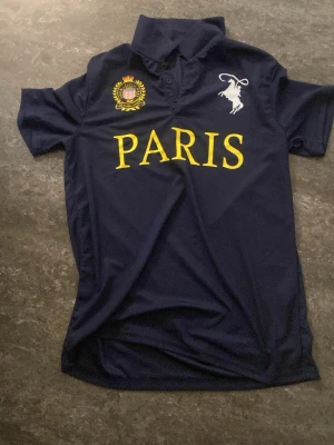 Mörkblå Paris pikétröja - Mörkblå pikétröja med gul text 'PARIS' på bröstet, broderad emblem och vit hästlogga. Klassisk krage och knappar framtill. Perfekt för en sportig och stilren look.