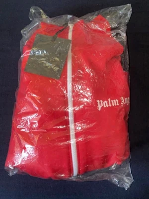 Röd Palm Angels dress - - Snygg röd dress från Palm Angels med vit dragkedja framtill och logga tryckt på bröstet. - Hoodien är tillverkad i mjukt material. Perfekt för dig som vill sticka ut med en ikonisk design.