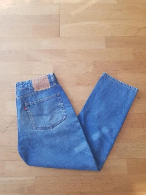 Levi's 505 blå raka jeans - Klassiska blå Levi's 505 jeans med rak passform och fem fickor. Jeansen har den ikoniska läderpatchen bak i midjan och röd Levi's-tagg på bakfickan. Tillverkade i slitstark bomullsdenim, perfekta för en avslappnad och tidlös stil.