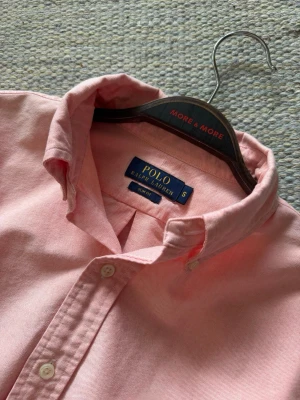Ljusrosa skjorta från Polo Ralph Lauren - Klassisk ljusrosa skjorta från Polo Ralph Lauren med button-down krage och broderad logga på bröstet. Skjortan har långa ärmar och knäppning framtill. Perfekt för en stilren look.