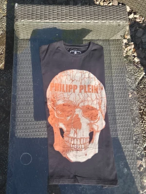Svart Philipp Plein t-shirt med dödskalle - Cool svart t-shirt från Philipp Plein med stor orange och vit dödskalle framtill och logga tryckt över pannan. Sen ett fett märke bak vid nacken och på sidan. Siiter riiiiktigt snyggt på , knappt använd varit inne på kemtvätt innan försäljning perfekt för dig som gillar edgy streetstyle. Värderad till 650-1300kr i detta skick är en m men sitter nästan som en s enligt Philipp Plein 