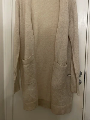 Beige lång kofta från Gina Tricot - Mysig beige lång kofta från Gina Tricot med två stora fickor framtill. Koftan är stickad och har en öppen front utan knappar, perfekt att slänga på sig över en t-shirt eller topp. Långärmad och i mjukt material.