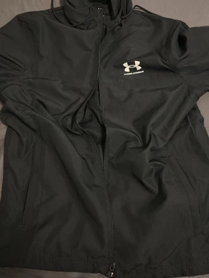 Svart vindjacka från Under Armour - Snygg svart vindjacka från Under Armour med dragkedja framtill och huva med dragsko. Jackan har en minimalistisk design med logga på bröstet och är tillverkad i lätt, vindtätt material. Perfekt för blåsiga dagar och sportiga outfits. Jackan är oanvänd!