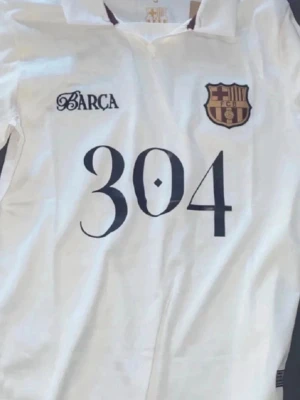 Vit FC Barcelona tröja Lamine Yamal - Vit sporttröja med FC Barcelona-logga och texten 'BARCA' på bröstet samt stort nummer 304. Baksidan har en stor grafisk illustration, klubbmärke och texten 'LAMINE YAMAL'. Perfekt för dig som älskar fotboll och vill sticka ut på planen.