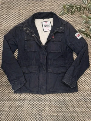 Mörkblå jacka från Superdry - Snygg mörkblå jacka från Superdry med fyra fickor framtill, dragkedja och knappar. Jackan har en patch på ena ärmen och mjukt foder upptill. Perfekt för kyliga dagar och har en klassisk, avslappnad look.