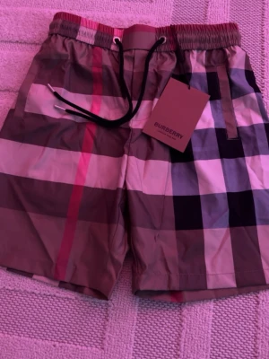 Burberry shorts - Snygga badbyxor från Burberry med klassiskt rutigt mönster i beige, svart, vitt och rött. Modellen har elastisk midja med svart dragsko och sidofickor. Perfekta för stranden eller poolen, med exklusiv känsla och ikonisk design.