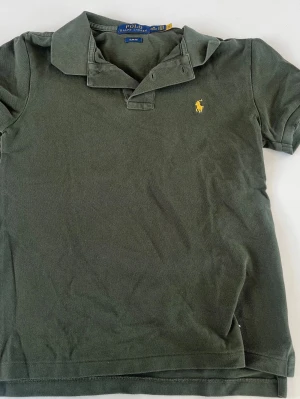Grön pikétröja från Polo Ralph Lauren - Klassisk grön pikétröja från Polo Ralph Lauren med korta ärmar och gul broderad logga på bröstet. Tröjan har krage och knappar upptill. Perfekt för en avslappnad och stilren look.