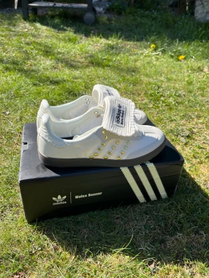 Adidas Wales Bonner white - Unika vita sneakers från Adidas x Wales Bonner med gulddetaljer och bred plös med broderad logga. Skorna har lågt skaft, klassisk gummisula och är tillverkade i läder med snygga sömmar och retrovibbar. Perfekta för dig som gillar stilrena och trendiga sneakers. Originalpris: 11 330 kr