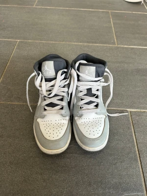Nike Air Jordan 1 Mid grå/vit -  Säljer ett par Nike Air Jordan 1 Mid sneakers i grått och vitt med svarta detaljer. Skorna har högt skaft, klassisk snörning och ikoniska Air Jordan-loggan på sidan. Perfekta för dig som gillar streetwear och vill ha en tidlös look. Om fler bilder önskas är det bara att höra av sig 💌
