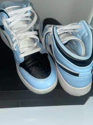 Jordan 1 Low UNC blå och svart - Snygga Nike Air Jordan 1 Low i UNC-färg med ljusblå, svart och vita paneler. Skorna har klassisk låg siluett, vit sula och svarta swoosh-detaljer. Ovandelen är i läder och de har vita skosnören. Perfekta för dig som gillar streetwear och sneakerskultur.