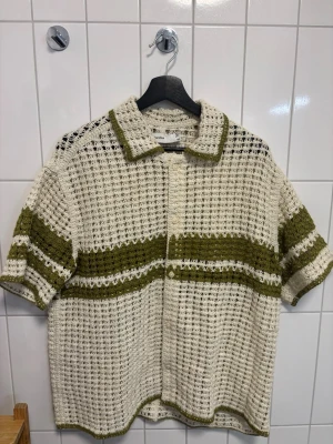 Resortskjorta från Bershka, beige/grön - Aldrig använd. Säljer en stickad skjorta från Bershka i beige med olivgröna ränder. Skjortan har korta ärmar, krage och knäppning framtill. Materialet är grovstickat och luftigt, perfekt för en avslappnad stil. Lite oversize modell.