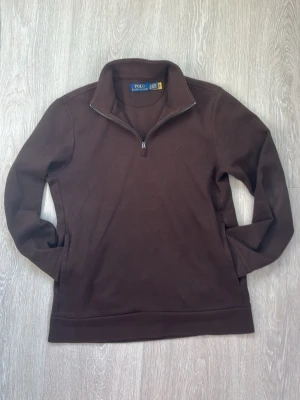 Brun halvzip från Polo Ralph Lauren - Snygg brun tröja från Polo Ralph Lauren med halv dragkedja och hög krage. Klassisk design med diskret broderad logga på bröstet. Perfekt för lager-på-lager och till chill outfits. Tillverkad i mjukt bomullsmaterial.