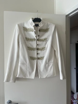 Vit kavaj med guldiga broderier - Stilren vit kavaj från Olivia London med ståkrage och dekorativa guldiga broderier i bladform på framsidan. Kavajen har knappar framtill och två fickor. Perfekt för dig som vill sticka ut med en unik och trendig look.