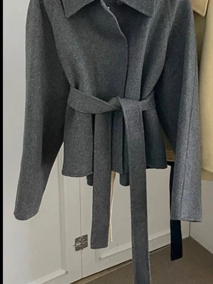 Grå kappa med bälte och bred krage - Stilren grå kappa med bred krage och knytskärp i midjan. Kappan har raka ärmar och två stora fickor framtill. Perfekt för dig som gillar en klassisk look med modern twist. Materialet känns mjukt och värmande, perfekt för kyligare dagar.