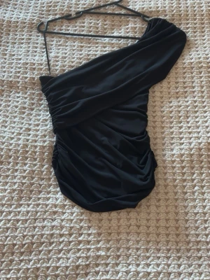 Svart oneshoulder topp - Snygg svart oneshoulder topp med draperad design och smal axelrem. Toppen har en tight passform och är perfekt för att skapa en trendig look. Materialet känns mjukt och följsamt mot huden.
