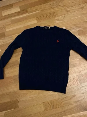 Mörkblå stickad tröja från Polo Ralph Lauren - Mörkblå stickad tröja från Polo Ralph Lauren med klassiskt kabelstickat mönster. Tröjan har rund halsringning och en liten röd broderad logotyp på bröstet. Perfekt för en stilren och avslappnad look.