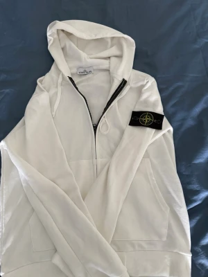 Vit StoneIsland Hoodie - En vit Stone Island-tröja i storlek Small kombinerar en lyxig känsla med en sportig, avskalad look. Den krispiga vitheten kontrasteras snyggt av den ikoniska kompass-loggan på vänster ärm, vilket ger plagget dess karaktäristiska identitet. Tröjan har en modern slim fit-passform som sitter kroppsnära och är tillverkad i kraftig, slitstark bomull av högsta kvalitet. Det är en tidlös klassiker som fungerar i alla sammanhang, så länge du kommer ihåg att knäppa loss patchen före tvätt för att bevara 