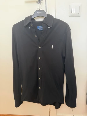 Svart skjorta från Polo Ralph Lauren - Klassisk svart skjorta från Polo Ralph Lauren med vit broderad logga på bröstet. Skjortan har långa ärmar, knappar framtill och en stilren krage. Perfekt för en clean och tidlös look.