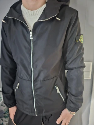 Svart vindjacka från Stone Island - Snygg svart vindjacka från Stone Island med huva och dragkedja framtill. Jackan har två snedställda fickor med dragkedja och den klassiska Stone Island-loggan på vänster ärm. Materialet är tunt och vindtätt, perfekt för blåsiga dagar.