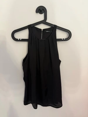 Svart halterneck topp från Gina Tricot - Säljer en svart, halterneck topp från Gina Tricot med rund halsringning och snygga veck framtill. Toppen har en lätt och luftig känsla, perfekt till jeans eller kjol. Dragkedja bak i nacken för en extra detalj.