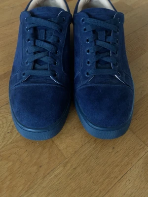 Blåa sneakers från Christian Louboutin - Snygga blåa sneakers i mocka från Christian Louboutin med klassisk röd sula och lågt skaft. Skorna har matchande blå snörning och en stilren design som sticker ut med den ikoniska röda undersidan.