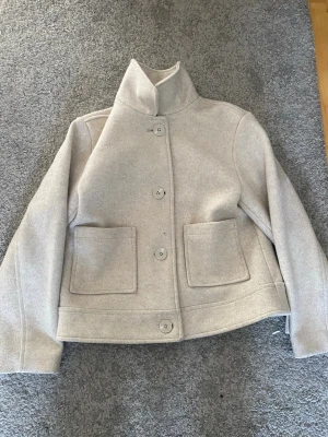 Beige ullkappa med hög krage - Stilren beige kappa i ullblandning med rak passform, hög krage och stora knappar framtill. Två rejäla fickor framtill ger en cool och praktisk look. Perfekt för kyliga dagar när du vill vara både varm och snygg.